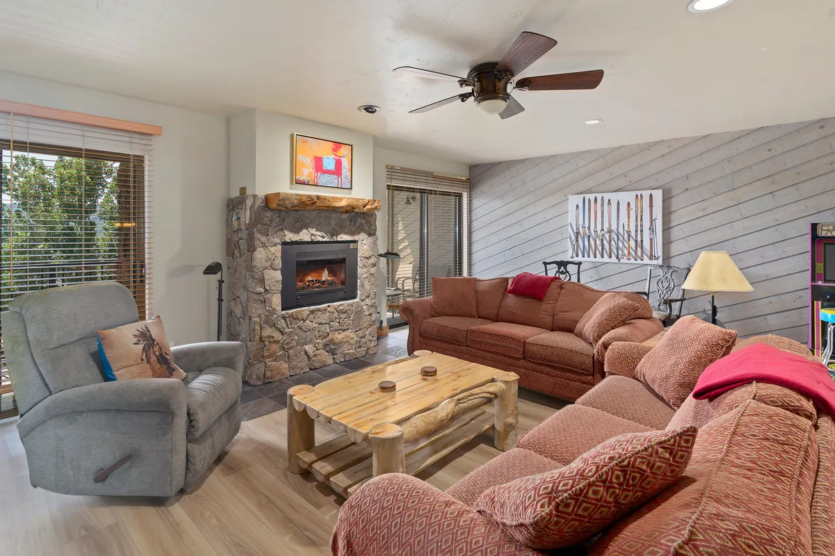 The image features a warm, inviting living room with a crackling fireplace, plush sofa seating arranged for conversation, a stylish coffee table, and tasteful mountain-inspired artwork adorning the walls. The space is designed for relaxation and gathering, perfect for reading, sharing stories, or unwinding after a day of adventure.