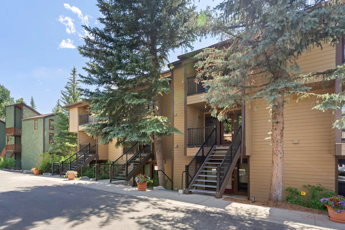 A welcoming exterior view showing wooden staircases, vibrant green foliage, and flower pots. The scene feels lively yet comfortable, suggesting community, approachable design, and opportunities for friends to gather before heading into town.