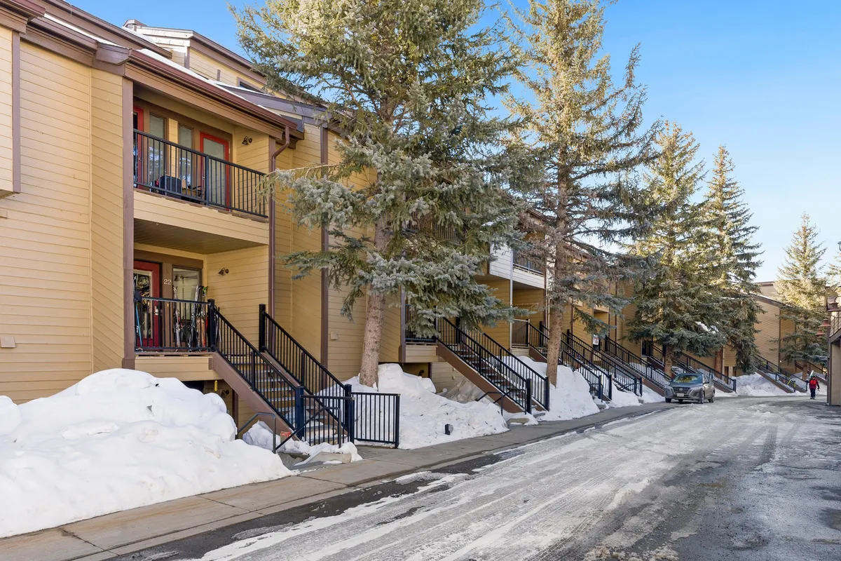 A welcoming exterior scene with wooden balconies, pine trees, a snow-covered courtyard, and a walkway leading through the property. It evokes a sense of community gatherings, winter sports, and exploration of the nearby woods.