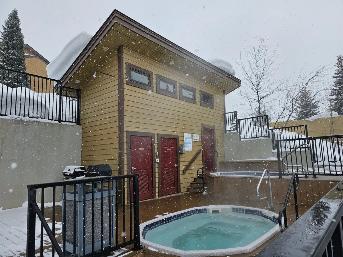 A communal hot tub surrounded by a snow-blanketed landscape and wooden buildings, steam rising invitingly above the water. The hot tub area is just steps from the accommodations, creating a cozy retreat after a day outdoors. The warm, inviting mood contrasts beautifully with the crisp winter setting.
