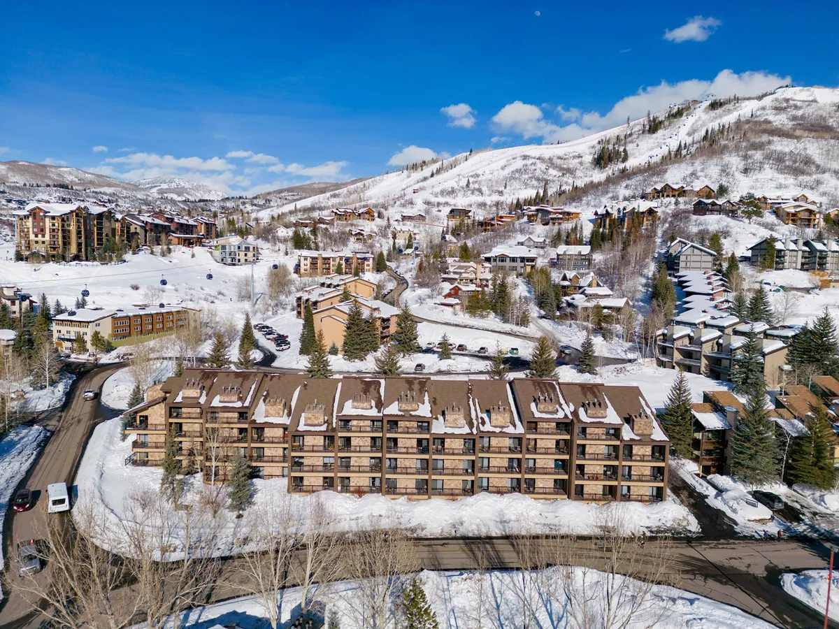 This image features snow-covered buildings nestled against a backdrop of majestic mountains under a clear blue sky. The scene evokes active winter fun, with visible ski tracks and a sense of adventure inherent in the setting. The overall mood is both adventurous and serene, reflecting the excitement of slope-side activities and the beauty of the natural landscape.