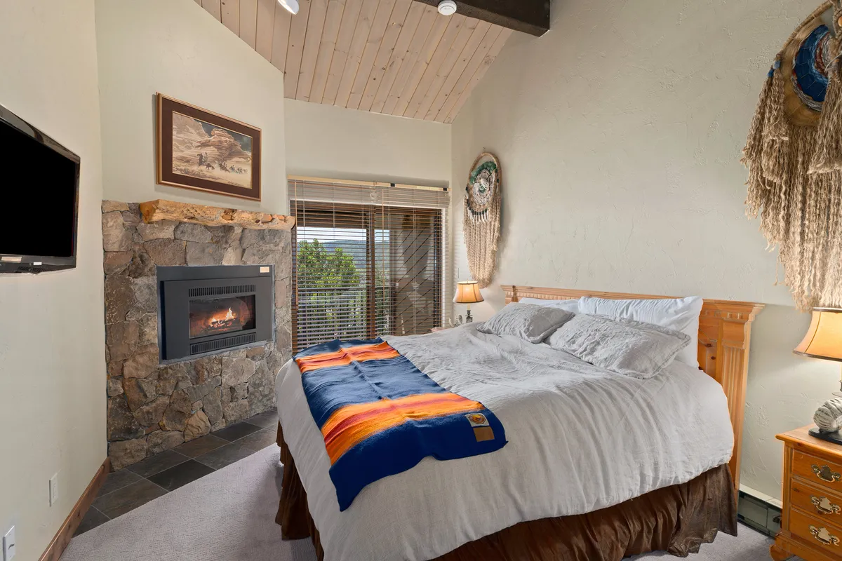 A stylish bedroom featuring a bed with a colorful blanket, a cozy fireplace, wooden furniture, and decorative wall hangings. The room exudes warmth and comfort, perfect for restful nights and quiet relaxation.